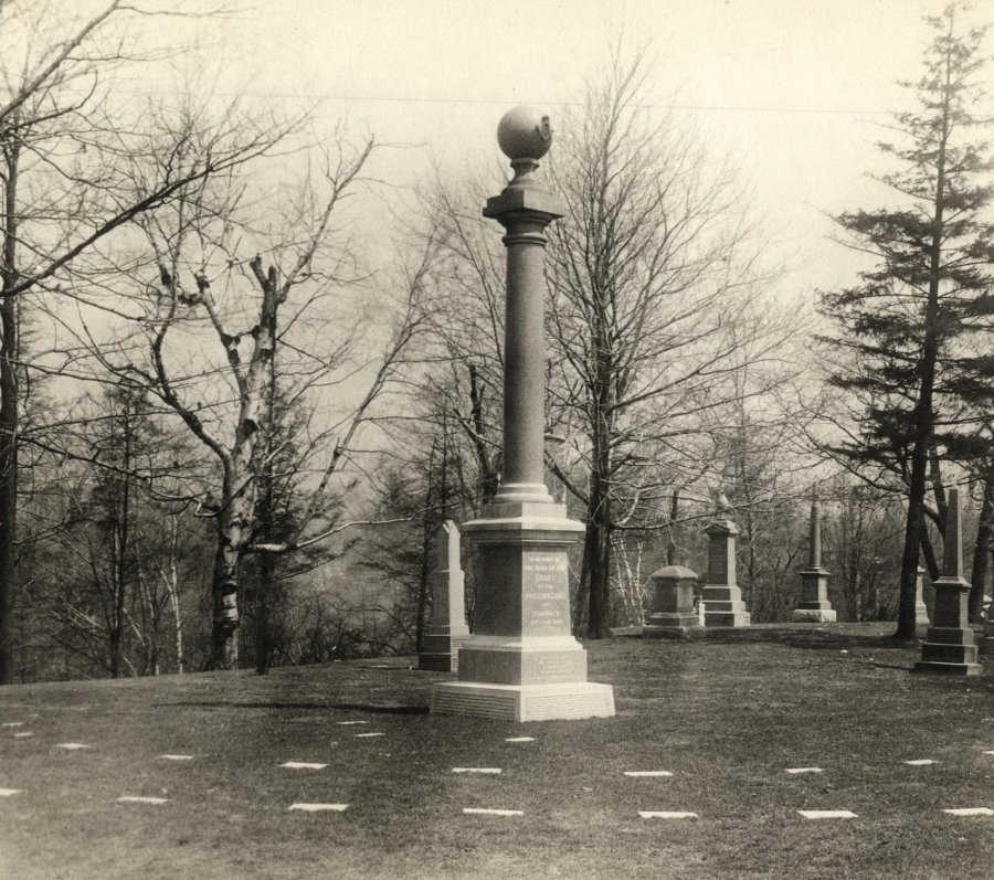 A black-and-white photograph of the Freemasons’ burial plot in a historic cemetery, featuring a tall stone column monument topped with a sphere in the foreground. Rows of smaller gravestones and monuments are arranged across a gently sloping grassy landscape, with leafless trees in the background, suggesting a formal memorial setting in late fall or early spring.