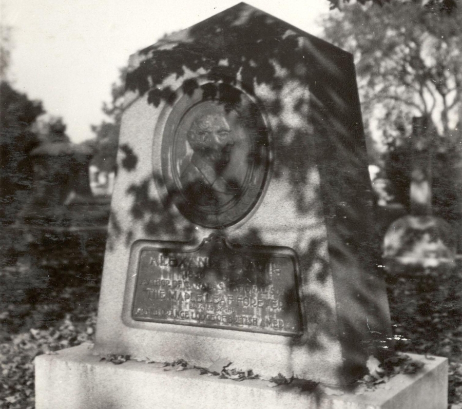 A black‑and‑white photograph of a stone grave monument standing outdoors among fallen leaves. The monument is tall with a sloped top and features an oval sculpted portrait relief of a man on its front. Beneath the portrait is an inscribed plaque, though the text is difficult to read due to shadows from nearby tree branches. Trees and other headstones are visible in the background.