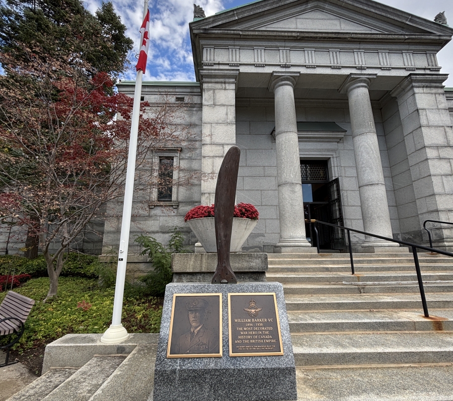 Outdoor photograph of a stone memorial set at the base of wide steps leading to a classical stone building with tall columns and a triangular pediment. In the foreground, a granite monument holds two bronze plaques and a vertical sculptural element shaped like an aircraft propeller. A Canadian flag stands on a pole to the left, with trees and landscaped greenery surrounding the area. The sky above is partly cloudy, and the scene conveys a formal, commemorative setting connected to history and remembrance.