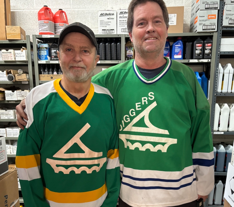 Two men standing side by side in a store aisle, wearing green hockey jerseys, with shelves of automotive supplies behind them.
