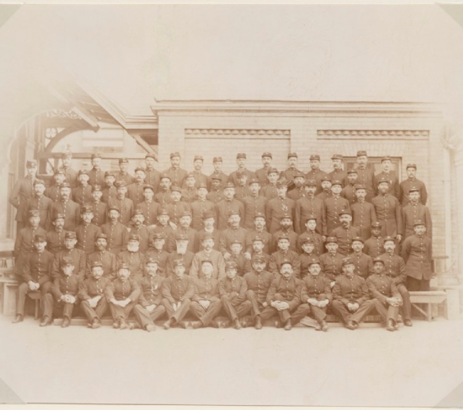 A large group of Toronto uniformed postal workers, including Albert Jackson, posed in several rows outside a brick building, with men seated and standing in formal arrangement, wearing matching caps and coats in a late‑19th‑century group portrait.