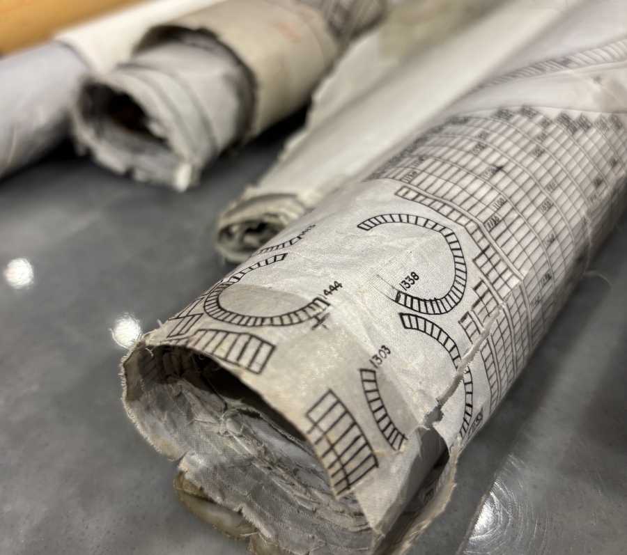 Rolled architectural or technical drawings resting on a table, showing printed measurement markings and curved line diagrams on aged, slightly torn paper.