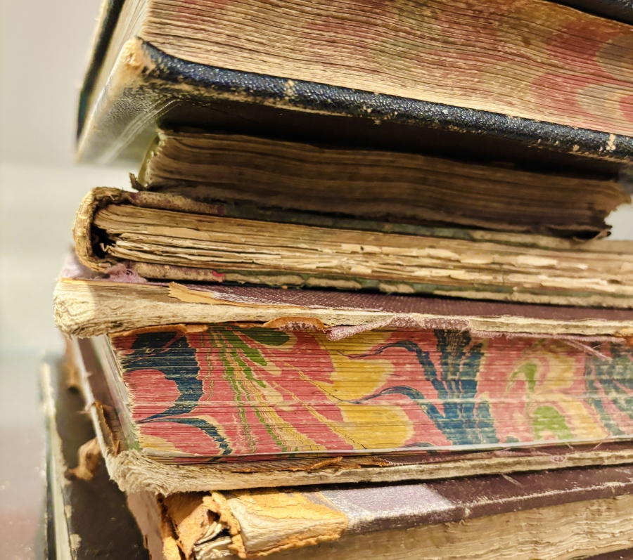 Stack of worn, old hardcover books with frayed edges and faded patterned covers, showing signs of heavy use and age, resting on a flat surface.