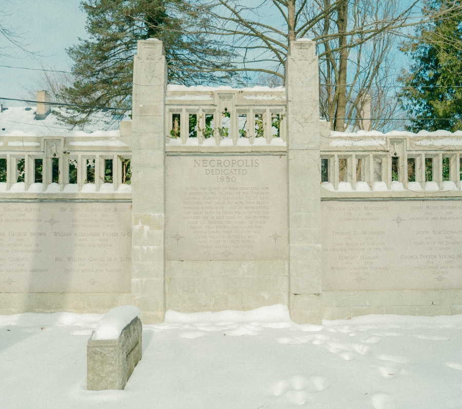 Snow‑covered stone dedication wall at the Toronto Necropolis, featuring engraved memorial panels framed by vertical pillars and decorative stonework. Trees rise behind the wall, and a low stone marker sits in the foreground within the winter landscape.