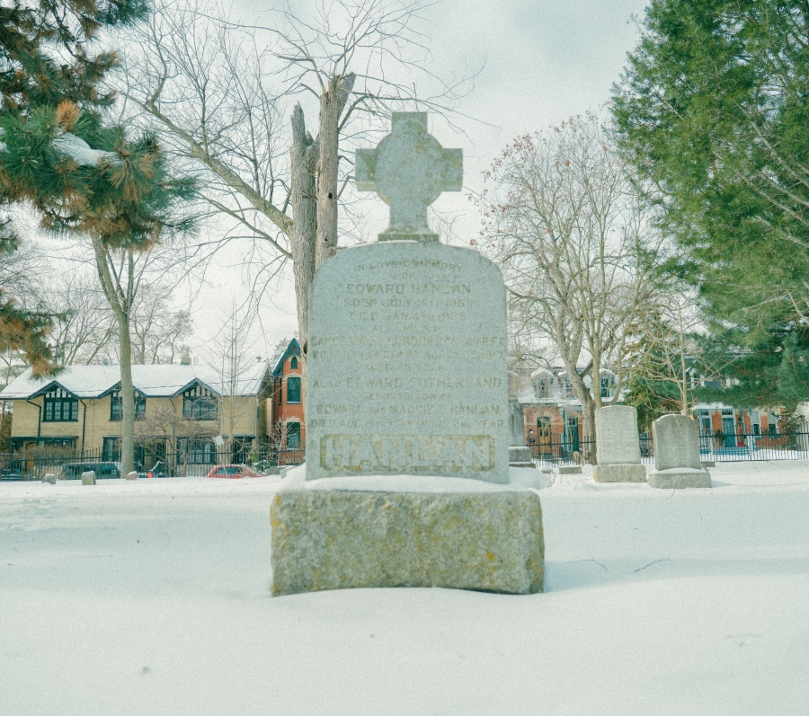 Snow‑covered cemetery scene featuring the grave marker of Ned Hanlan, a tall stone headstone topped with a cross and set on a rectangular base. The monument stands among trees and surrounding grave markers, with residential buildings visible in the background during winter.