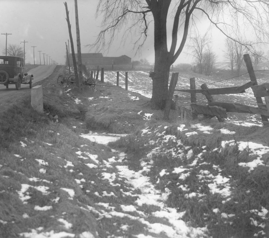 A black-and-white photograph of an undeveloped rural roadside in winter, with patches of snow scattered across uneven ground in the foreground. A dirt road runs along the left side of the image, with an early automobile parked near a line of utility poles. Wooden fence posts, a leafless tree, and open fields extend into the distance, with farm buildings faintly visible on the horizon.
