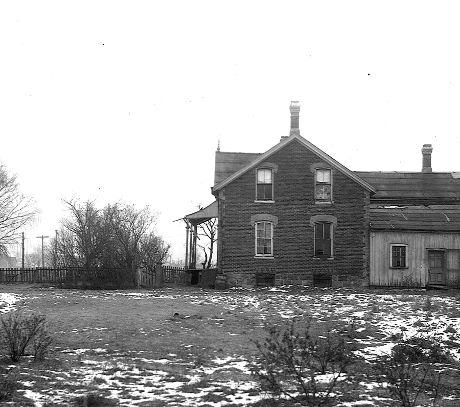 A black-and-white photograph of a rural brick farmhouse with a gabled roof and a small wooden addition on one side, set on an open field with patches of snow on the ground. Leafless trees, a fence line, and utility poles surround the property, suggesting a winter scene in an early 20th-century rural setting.