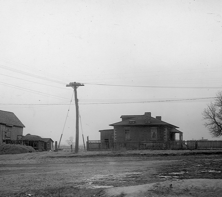 A black-and-white photograph of a small rural settlement showing a one-storey house with a hip roof and front porch enclosed by a wooden fence, alongside a large wooden barn and outbuildings. A dirt road runs across the foreground, with utility poles and overhead wires cutting across the scene, leafless trees nearby, and patches of snow on the ground, suggesting a winter landscape from the early 20th century.
