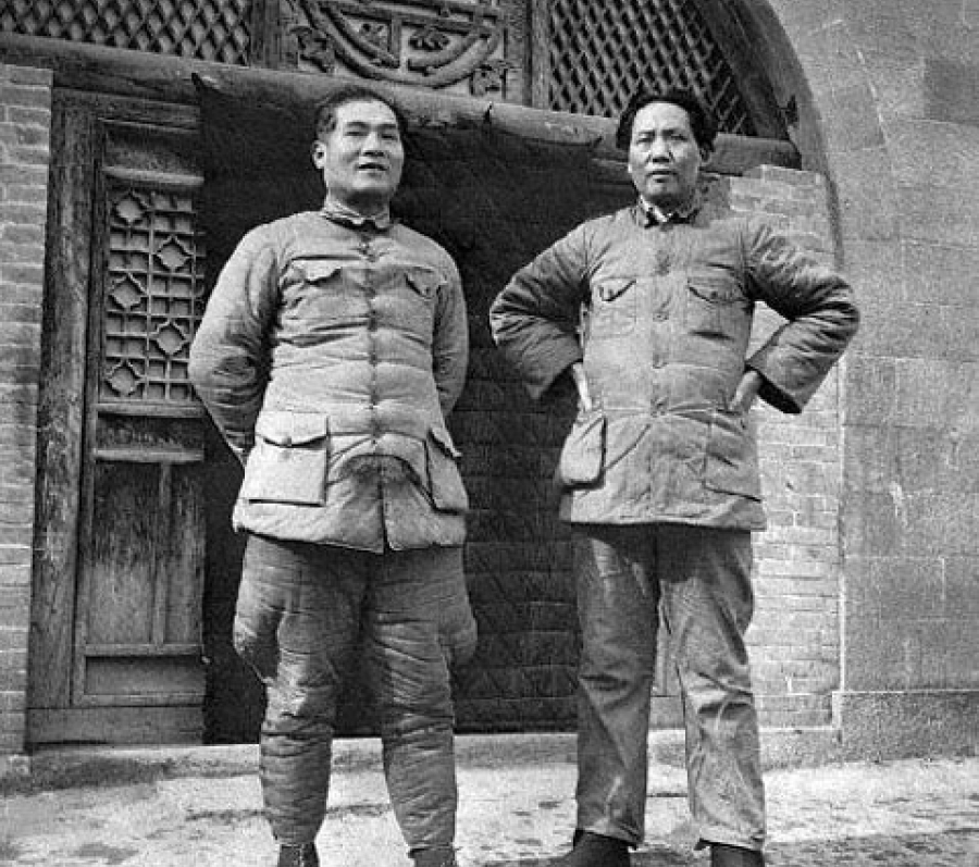 Black‑and‑white photograph of two people wearing padded military jackets, standing side by side in front of a stone building with a carved emblem above the doorway. Both figures stand confidently with hands positioned at their sides or on their hips.