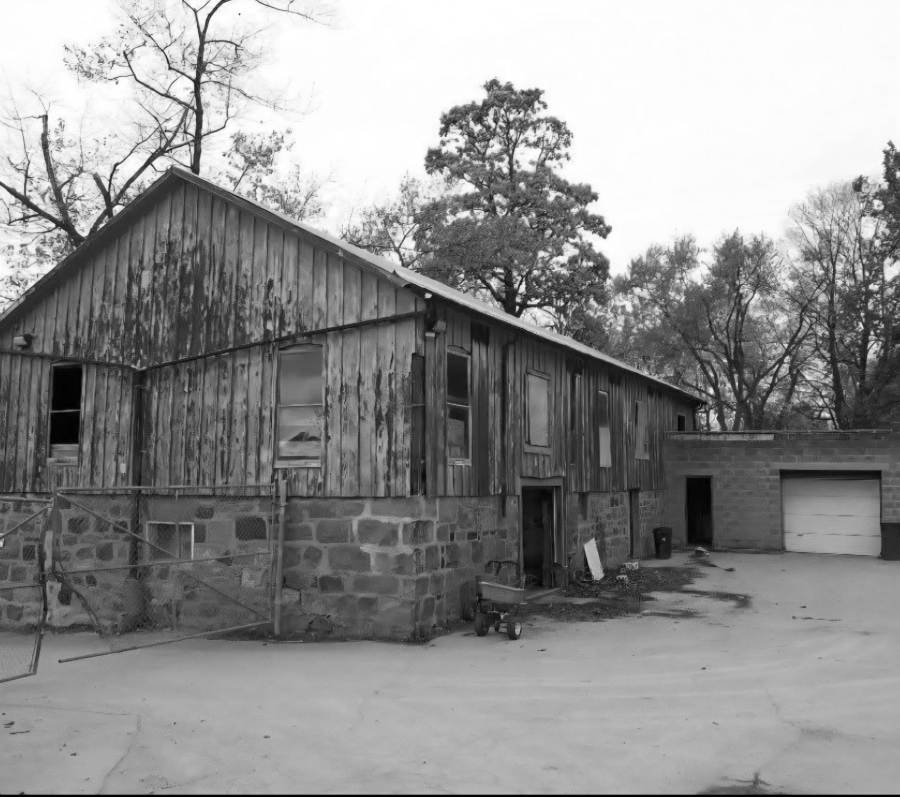 Black and white photograph of the barn structure in the Pine Hills Cemetery maintenance yard. There are tall trees behind, some are bare and others are coniferous.