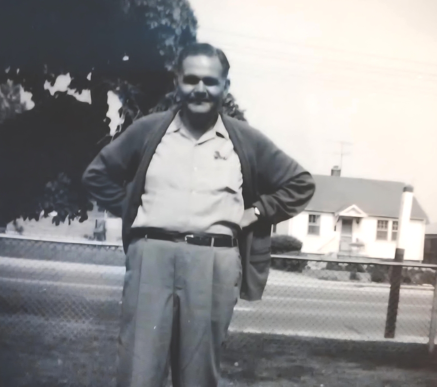 Black and white photo of Mel Chandler standing with his hands on his hips. He is wearing a light-coloured button-down dress shirt, belted dress pants, and a cardigan. Behind him is a large leafy tree and a house across the road. Behind him are the gates to Pine Hills Cemetery, at the Kennedy side exit.