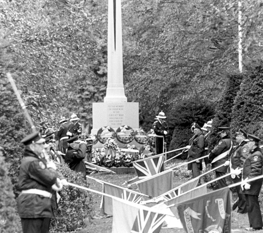 Black-and-white photo of a ceremonial gathering at a memorial cross, with uniformed participants standing in formation and flags displayed in front of the monument, surrounded by trees.