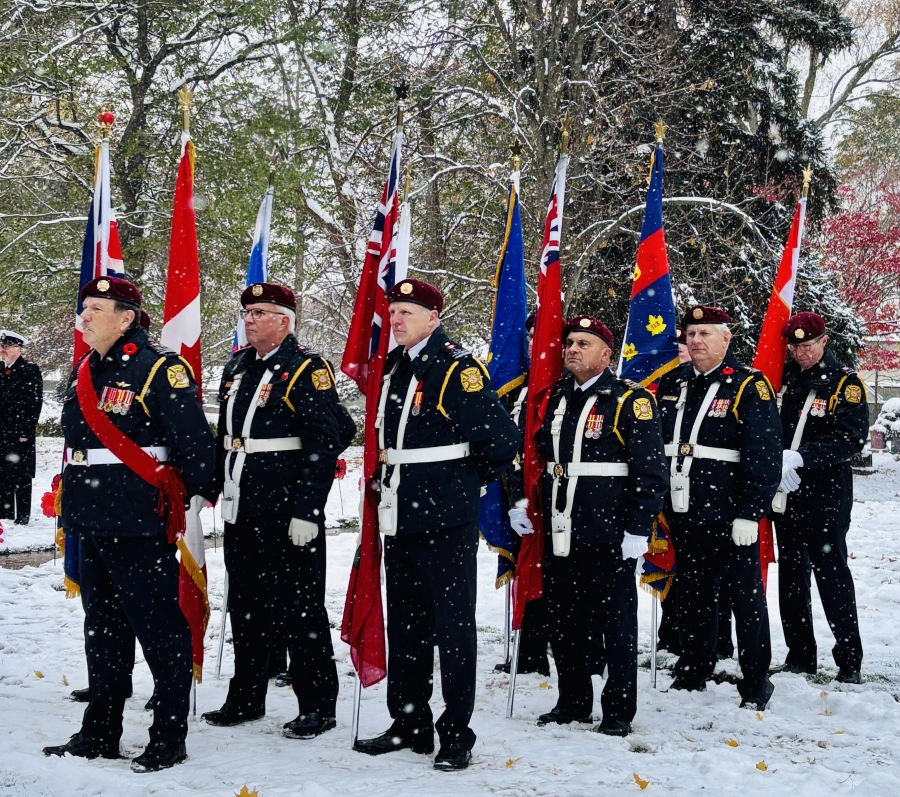 Six servicemen carrying flags, standing with their arms to the side. There is snow on the ground and the trees in the background have snow on their leaves and needles.