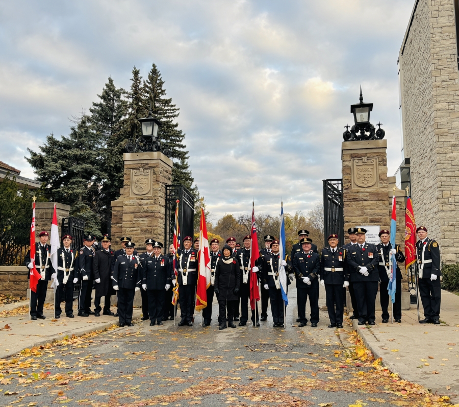 About 25 people gather between two stone pillars at the gate of Prospect Cemetery. There are a few flags held by members of HCMS York. The mayor of Toronto, Olivia Chow is in the centre of the photo. There are yellow leaves on the road leading into the cemetery, and the sky is cloudy with some blue sky peeking through.