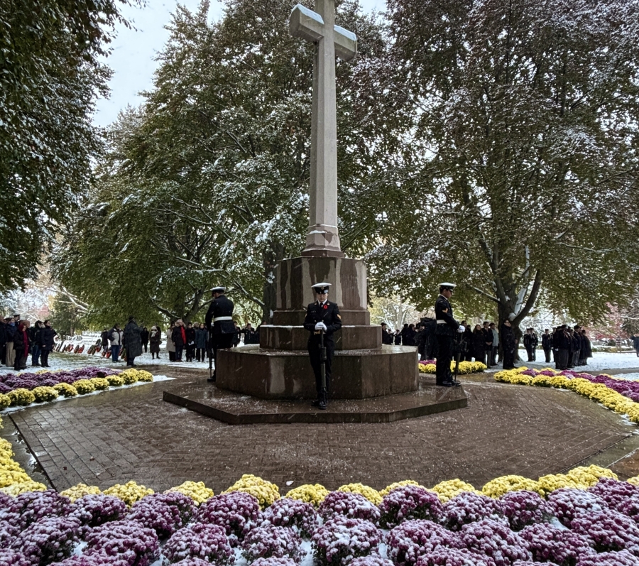 A snow-dusted veterans memorial cross stands at the center of a circular garden filled with purple and yellow flowers, surrounded by tall trees. Uniformed guards stand watch as people gather quietly in the background, creating a solemn, reflective atmosphere.
