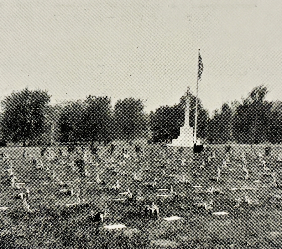 Historic black-and-white photograph of a veterans’ burial plot with rows of small grave markers spread across a grassy field, centered on a tall Cross of Sacrifice with a flag. Mature trees line the background