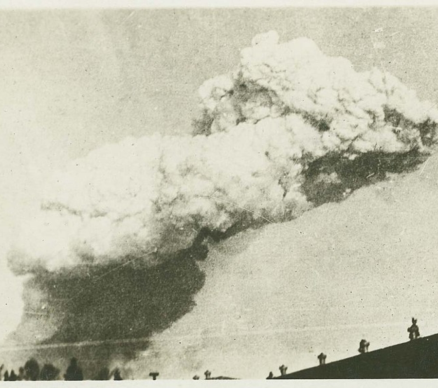 Historic black‑and‑white photograph showing a massive explosion cloud rising into the sky above buildings and rooftops. The cloud billows outward in dense layers, dominating the horizon while silhouetted structures and figures appear along the lower edge of the image.