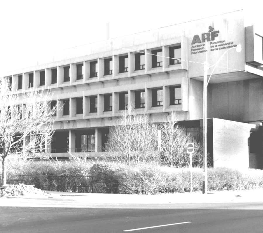 A black‑and‑white exterior view of the Addictions Research Foundation where Eleanor worked as a therapist. It is a mid‑century institutional building with a concrete façade and rows of rectangular windows, set back from a city street with bare trees and low shrubs in the foreground. The photo was taken in the 1990s.