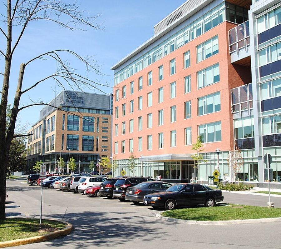 Exterior view of a modern multi‑storey institutional building with red brick and glass façades, large windows, and a stepped design, fronted by a parking lot with cars, young trees, and pedestrian walkways under a clear blue sky.