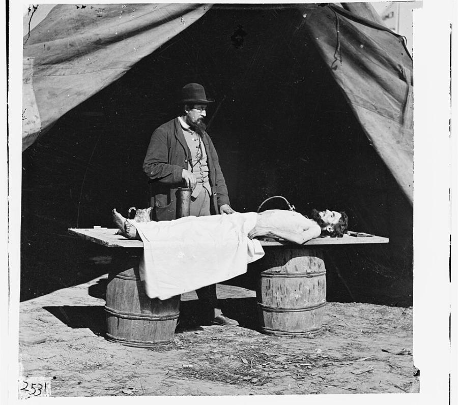 Black‑and‑white historical photograph showing an embalming surgeon standing beside a covered body laid on a wooden table supported by barrels, set outdoors beneath a canvas tent. Medical equipment and containers rest on the table, and the ground beneath is bare earth, reflecting a field mortuary setting.