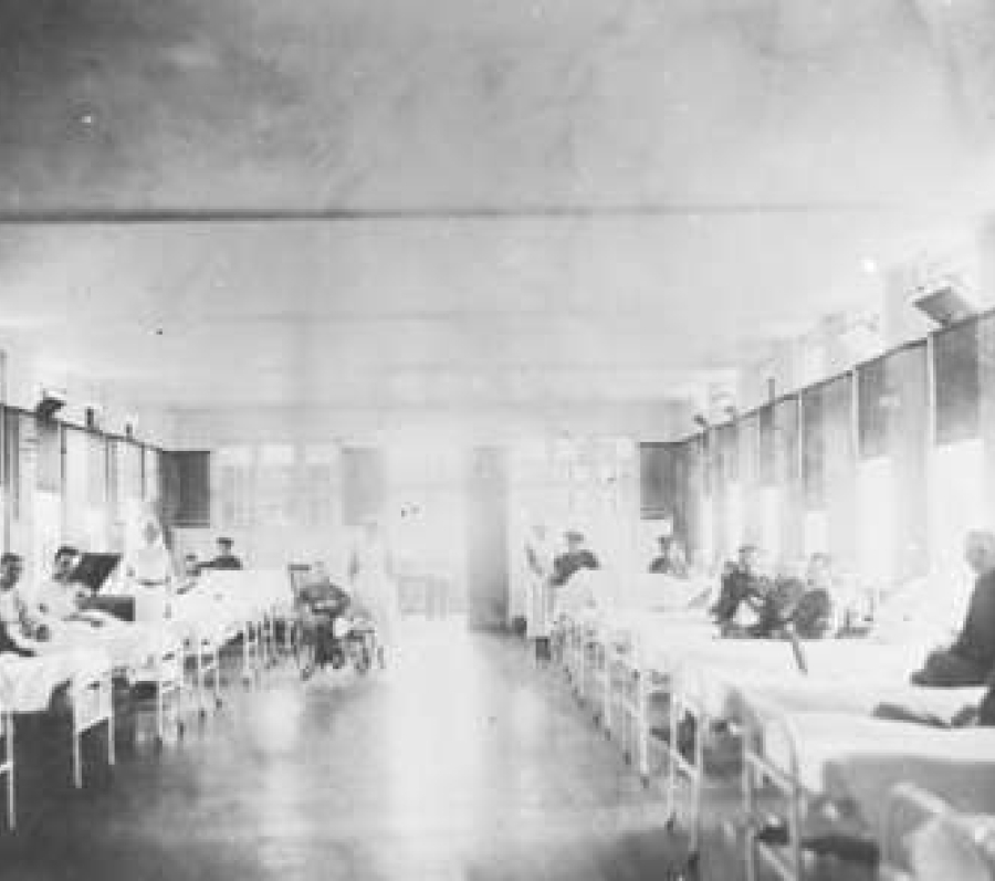 Black‑and‑white photograph of a long hospital ward with rows of metal‑framed beds on either side, occupied by recovering soldiers, with nurses attending patients beneath large windows lining the room.