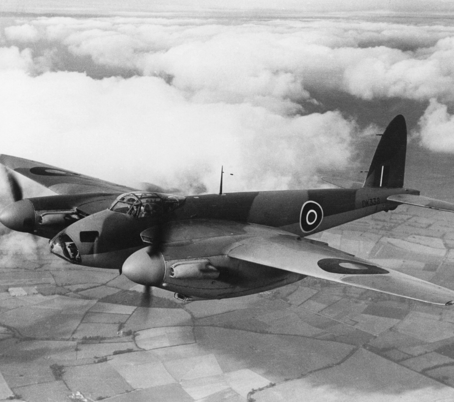 Black‑and‑white photograph of a military aircraft flying above patchwork farmland and clouds. The twin‑engine plane bears Royal Air Force roundels on its wings and fuselage and is shown in flight from a side angle, with clouds stretching across the horizon below.