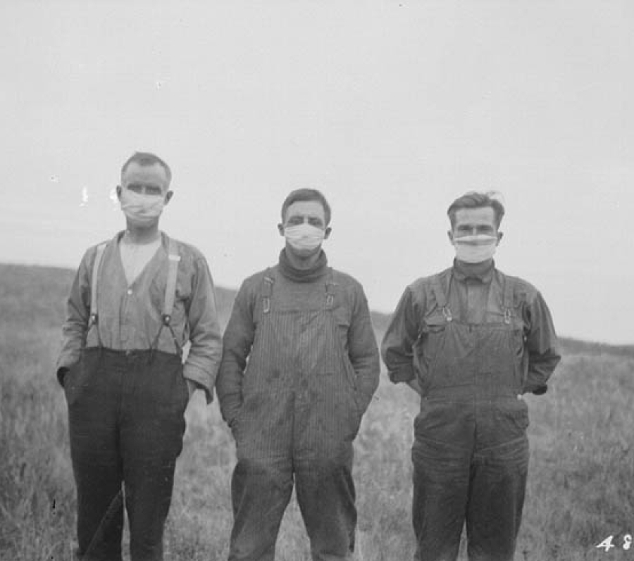Black-and-white photograph of three young people standing side by side in a grassy field. They are wearing heavy work clothes with long sleeves and overalls, suggesting farm or manual labor. All three of them are wearing medical masks. The field slopes gently behind them, with open land and sky in the background. The scene has a stark, historical feel, with simple clothing and a rural setting.