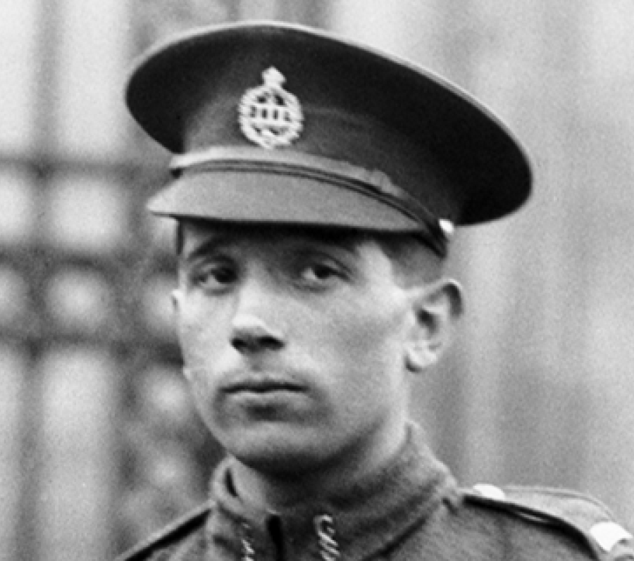 A historical black‑and‑white photograph of a uniformed military figure wearing a peaked cap with an insignia. The person is dressed in a formal service uniform, standing outdoors in front of a softly blurred background with architectural elements.