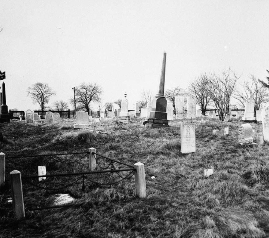 Black and white photo of a burial ground. There are some trees in the background, and it appears to be late autumn. 