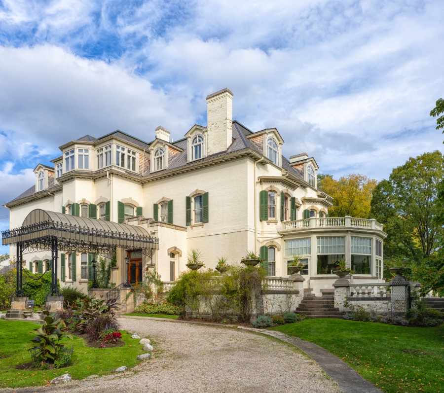 Exterior view of Spadina House, a historic two‑and‑a‑half‑storey mansion with cream‑coloured walls, green shutters, and a mansard roof, featuring a covered entrance, bay windows, and landscaped gardens with a curved driveway and mature trees under a partly cloudy sky.