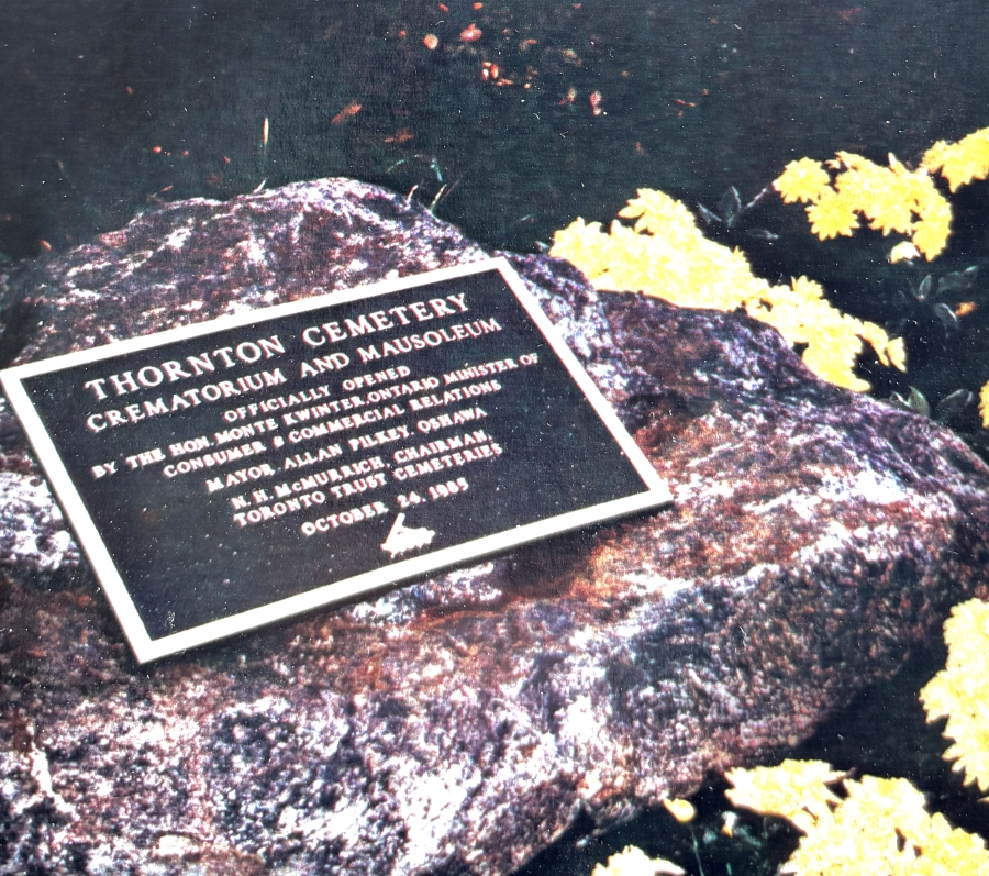 A commemorative plaque mounted on a large rock at Thornton Cemetery marks the opening of the crematorium and mausoleum, surrounded by yellow flowers and fallen leaves outdoors.
