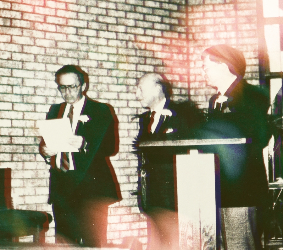 Three men in formal suits stand at a podium in front of a brick wall. A letter from Ontario Premier David Peterson is being read by the Honourable Monte Kwinter, just prior to being sealed in a time capsule. To his right is Norman McMurrich, and next to him is Oshawa Mayor Alan Pilkey. They are inside the building at Thornton Cemetery. There is a stained glass window behind them on the right of the frame.