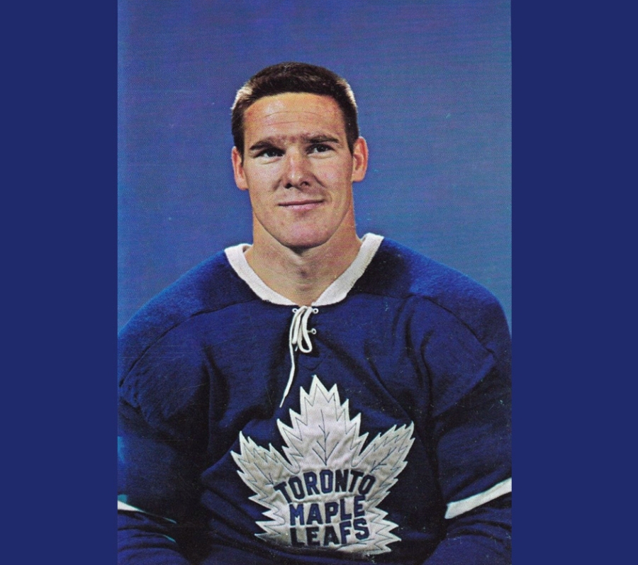 Tim Horton wearing a vintage Toronto Maple Leafs jersey poses in front of a solid blue background. The player is shown from the waist up, dressed in a blue sweater with a large white maple leaf logo and lace-up collar.