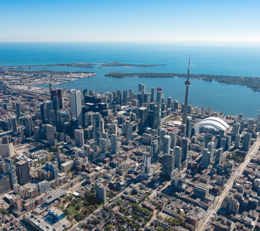 Aerial view of downtown Toronto on a clear day, showing a dense cluster of high-rise buildings, the CN Tower, the Rogers Centre, and Lake Ontario stretching out in the background.
