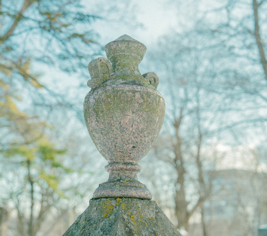 Close‑up view of a carved stone urn atop a grave monument, weathered with patches of lichen. Leafless trees and soft daylight blur into the background, creating a calm, reflective cemetery setting.