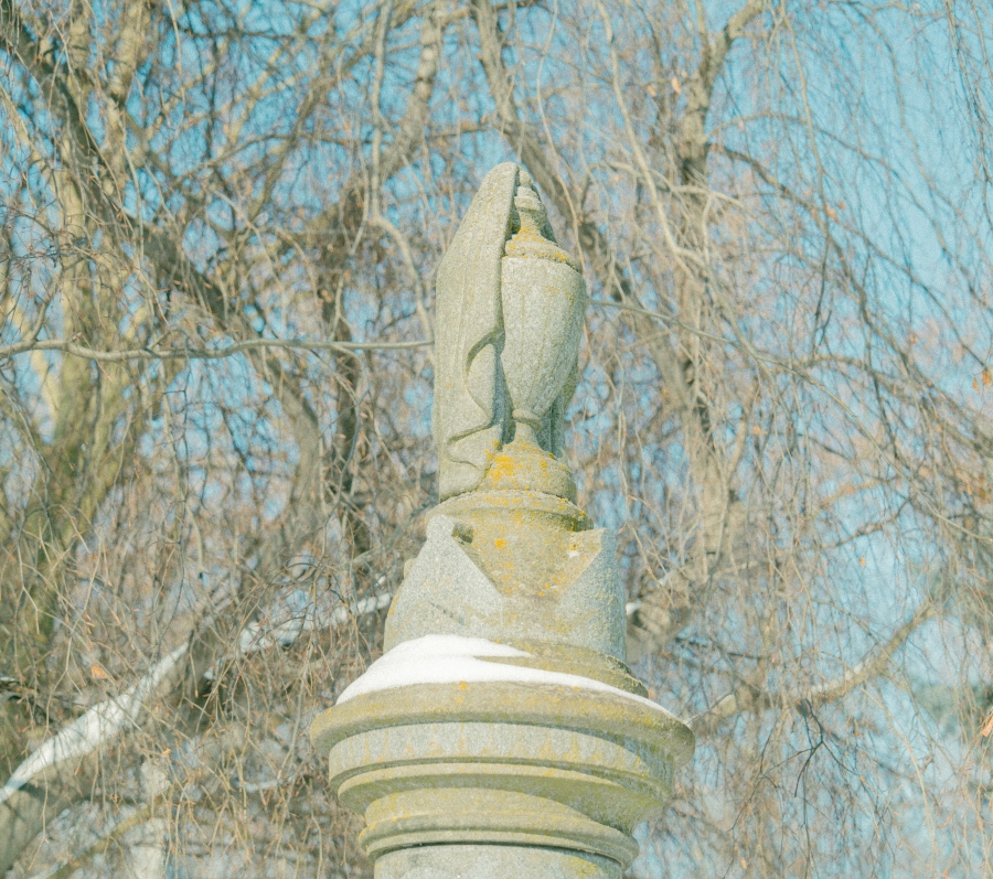 Close‑up view of a carved stone urn atop a grave monument, weathered with patches of lichen. Leafless trees and soft daylight blur into the background, creating a calm, reflective cemetery setting.