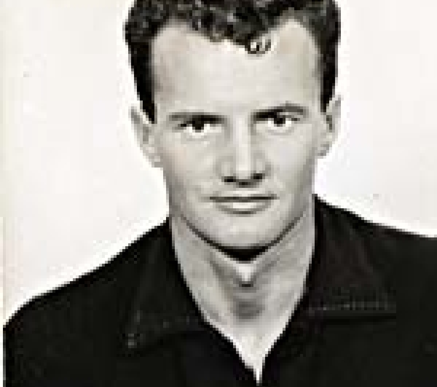 A black and white photo of Walter Grealis wearing a black button up shirt. He has short styled hair with a curly lock in the front. 