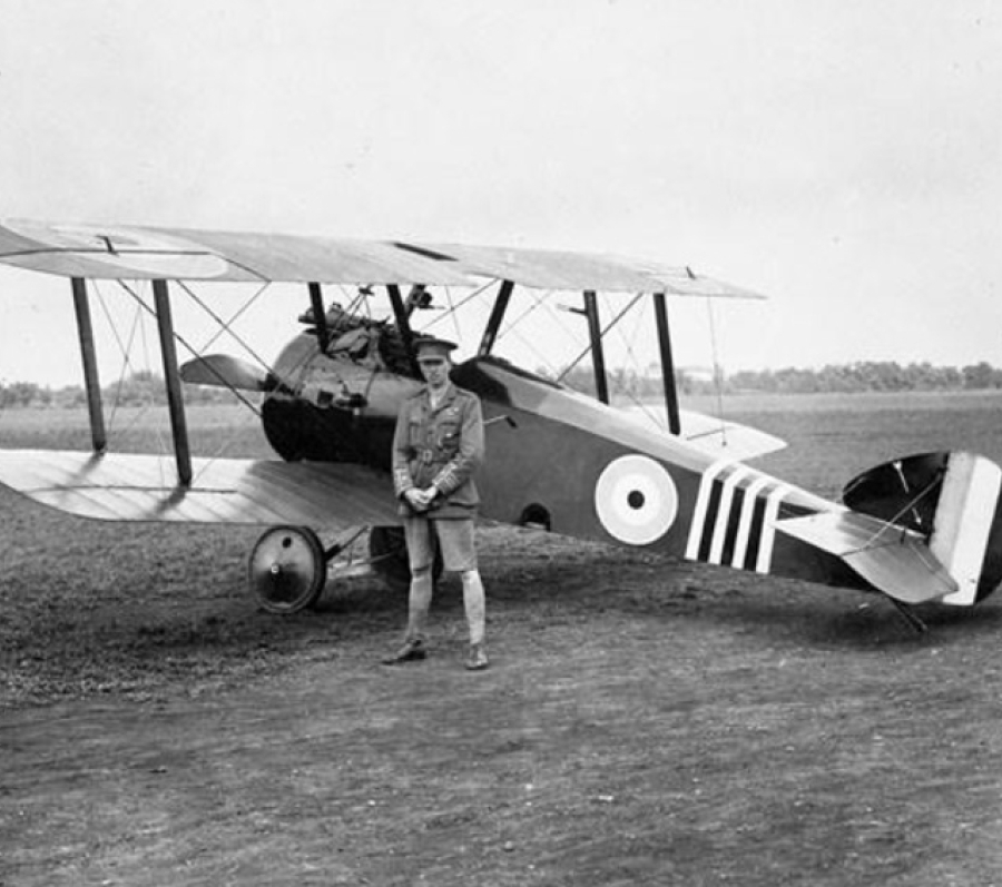 A vintage black‑and‑white photograph of a World War I–era biplane parked on an open airfield. The aircraft has roundel markings on its fuselage and bold striped patterns on the tail. A uniformed pilot stands in front of the plane, hands clasped, looking toward the camera. The background shows flat terrain with trees along the horizon.