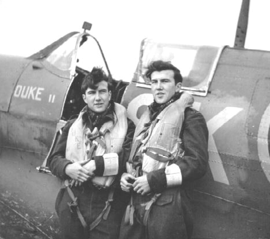 Black-and-white photograph of the identical twins Bruce and Duke Warren standing together beside a fighter aircraft marked “DUKE II.” Both wear flight jackets and life vests, with the cockpit and nose of the plane visible behind them, suggesting preparation for or return from a flight.