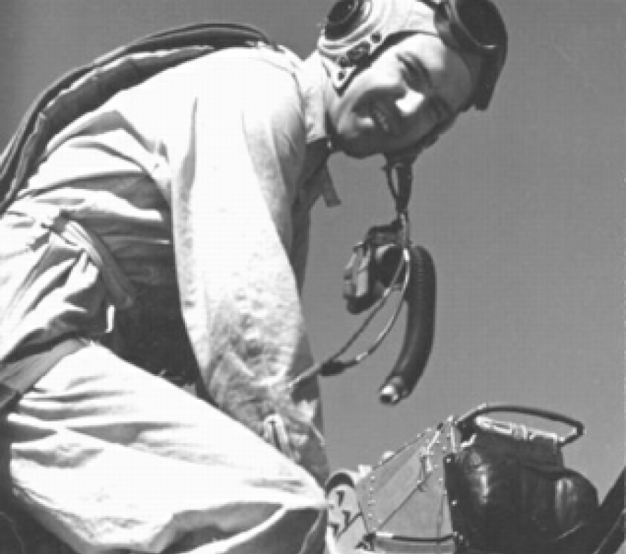 Black-and-white photograph of Bruce Warren wearing a flight suit, helmet, and goggles, crouched beside an aircraft while working with equipment on the fuselage. The angle is close and slightly below, emphasizing the technical details of the gear and the aircraft surface.