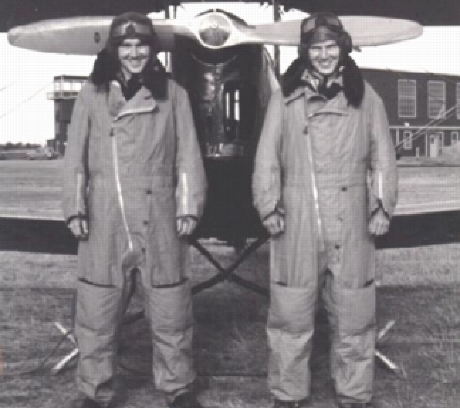Black-and-white photograph of the twin aircrew members, Bruce and Duke Warren, standing side by side on a grassy airfield in front of a small military aircraft. They are wearing heavy flight suits and boots. They are smiling and posing in front of the plane with hangar buildings visible in the background.