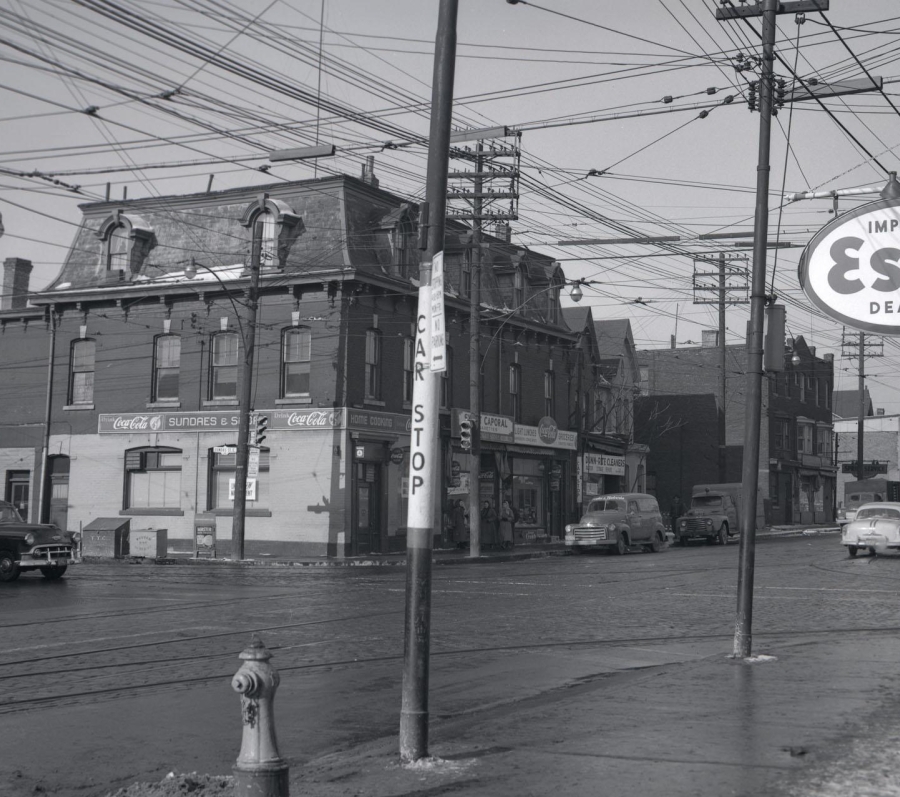 Black‑and‑white photograph of a city street intersection in 1957, showing brick commercial buildings, overhead streetcar wires, parked cars, and storefront signage. An “Imperial Esso Dealer” sign stands on the sidewalk, with utility poles, a fire hydrant, and early automobiles visible along Parliament Street in Toronto.