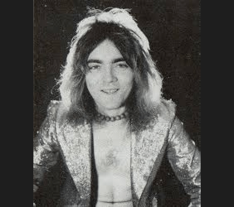 Black‑and‑white studio portrait of John Rutsey with long hair, wearing a patterned jacket over a light‑coloured shirt, posed against a dark background.