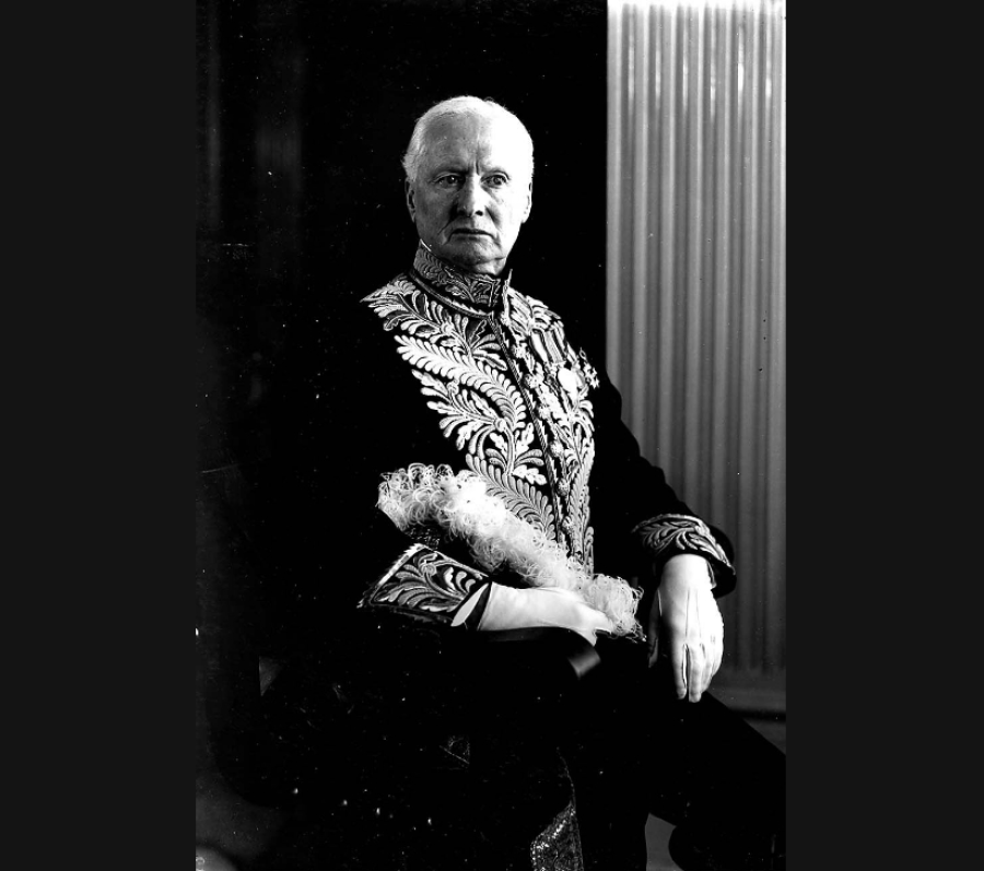 Black‑and‑white formal studio portrait of a person seated and wearing an ornate, embroidered ceremonial jacket with decorative braiding and medals. The individual rests one arm on a chair, with a feathered accessory across their lap, posed against a dark backdrop with a textured column visible to one side.