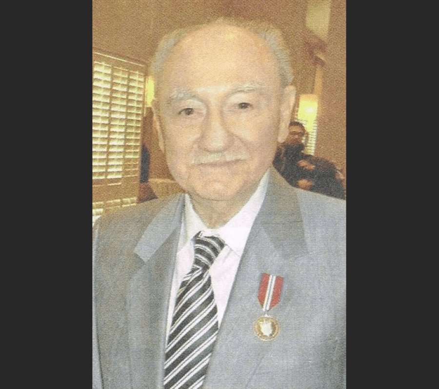 Formal portrait of a person wearing a light‑coloured suit and striped tie, with a military or service medal pinned to the left lapel. The photograph appears to be taken indoors, with window blinds and seated figures visible in the softly lit background.