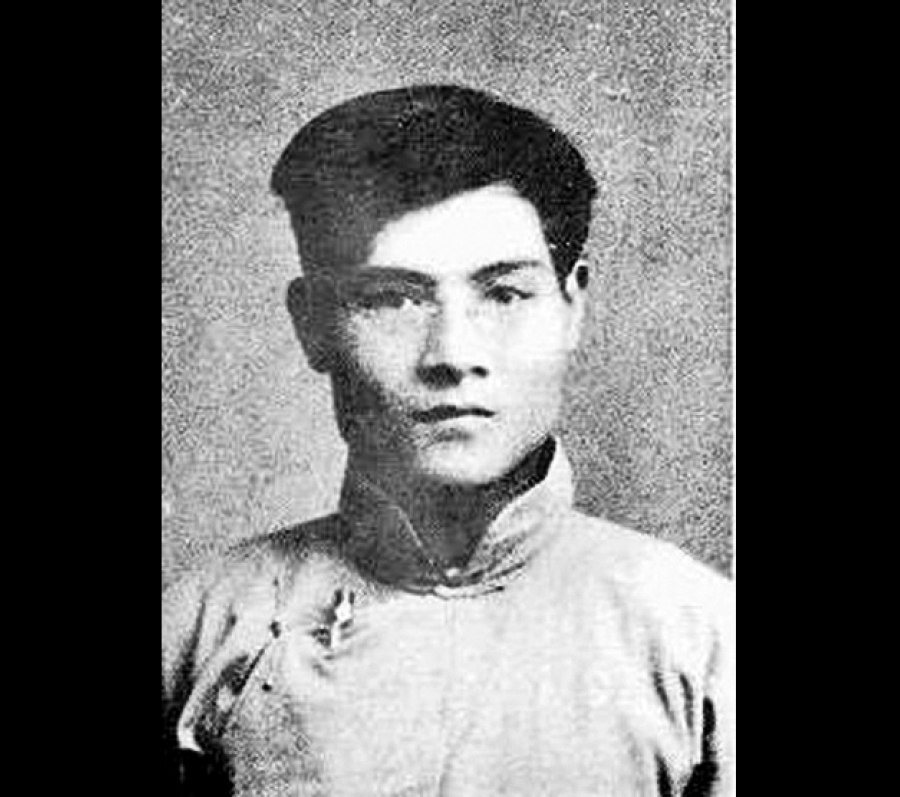 Black‑and‑white portrait of a person wearing a cap and a simple, long‑sleeved shirt, photographed from the chest up against a plain textured background.