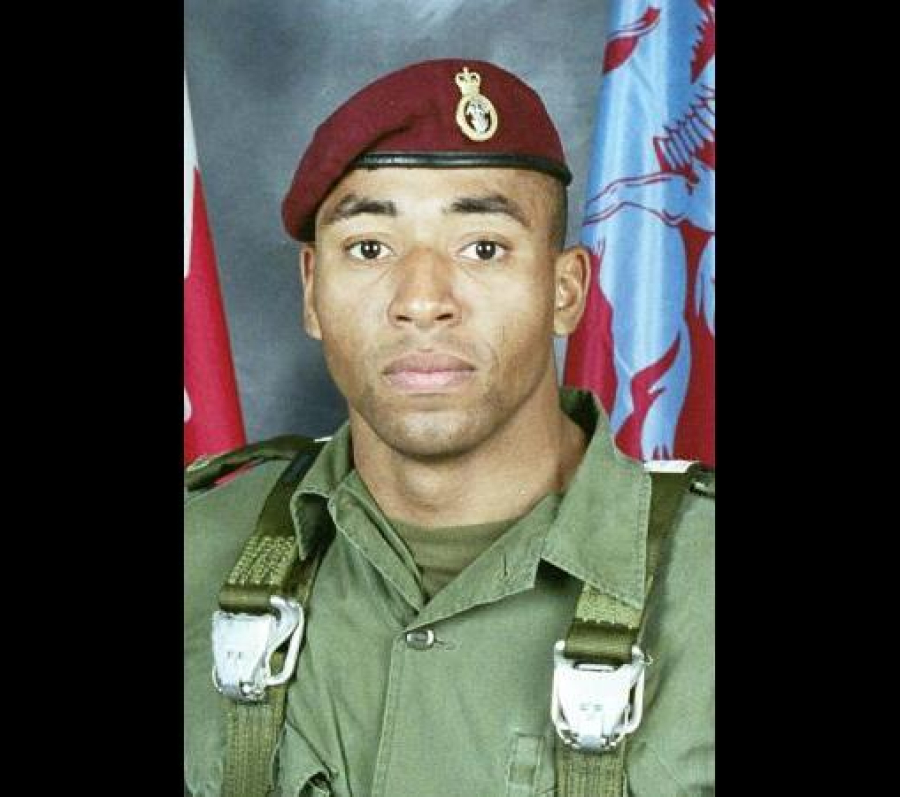 Portrait of Corporal Ainsworth Dyer in military uniform, wearing a maroon beret and standing in front of flags, photographed from the chest up.