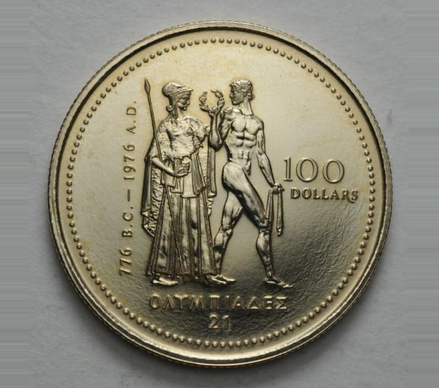 A close-up photograph of a gold commemorative coin featuring two classical figures standing in the center—one draped and holding a staff, the other nude and athletic, holding a wreath. The inscriptions include “776 B.C. – 1976 A.D.” along the left side, “100 DOLLARS” on the right, and Greek lettering along the bottom. The coin rests on a light, textured background.
