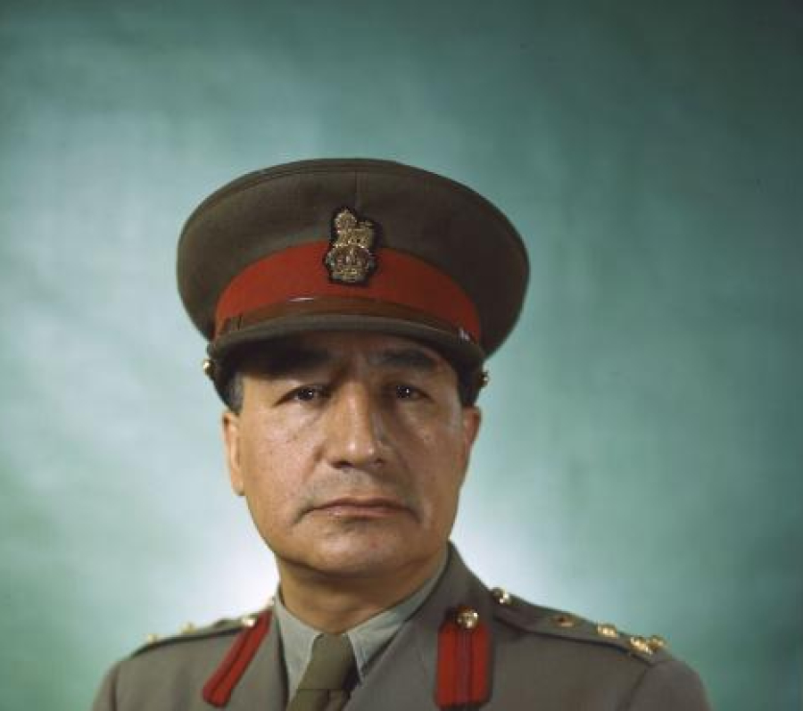 Studio portrait of a military officer wearing a formal uniform with a peaked cap, medal ribbons, and aviation wings, posed against a plain teal backdrop and shown from the waist up.