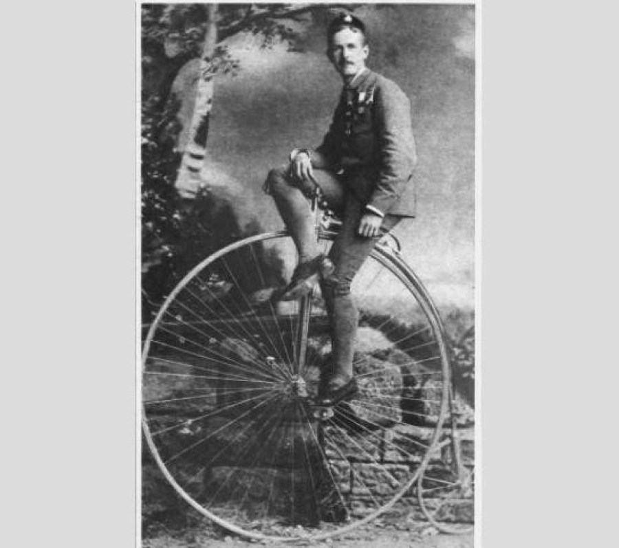 Historic black‑and‑white photograph of a man wearing a suit and tie, posed seated on a large high‑wheel bicycle in an outdoor studio setting with a painted backdrop of trees and foliage.